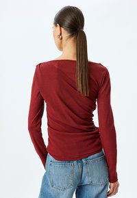 Bordeauxrotes Langarmoberteil mit drapiertem Design, weichem Material und weitem Ausschnitt, kombiniert mit hellblauen Jeans.