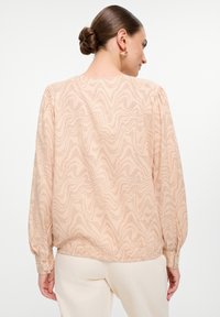 Blusa color pesca chiaro con un motivo ondulato, maniche lunghe e orlo arricciato. Presenta polsini con bottone e un tessuto liscio e strutturato.