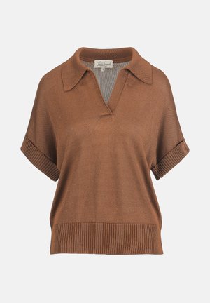 Femeie purtând un top tricotat maro cu mâneci scurte, pantaloni maro și albi cu model, sandale maro, ținând o poșetă maro și purtând brățări groase.