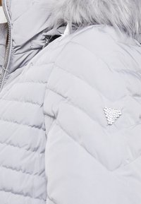 Veste d'hiver gris clair matelassée avec une capuche bordée de fourrure et un groupe triangulaire de petites perles brillantes sur la manche.