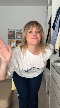 Balta T-krekls ar "PALM SPRINGS" druku, apvienots ar tumši ziliem biksēm. Fons ietver plauktus ar karājošiem apģērbiem un žurnālu vākiem.