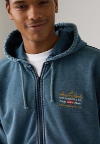 Blå hettegenser med glidelås laget av teksturert stoff, med brodert merkevare i gull og hvitt. Hette med snor og frontglidelås.