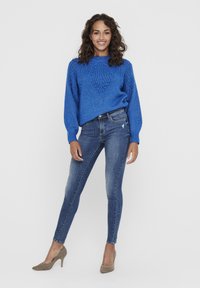 Blue knit sweater with textured pattern, paired with fitted blue jeans. Model wears light gray high heels. Plain white background.