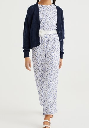 Tyttö, joka on pukeutunut valkoiseen ja siniseen kukkakuosiseksi haalariin, jossa on valkoinen vyö, tummansiniseen neuleeseen ja valkoisiin sandaaleihin, kävelee eteenpäin.