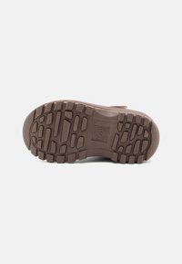 Suela de zapato de goma marrón que presenta una superficie texturizada con hendiduras y patrones profundos para tracción. Marcada con un logo circular en el centro.