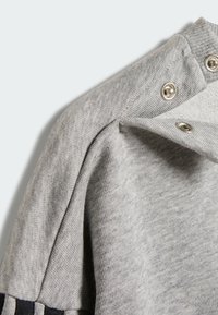 Grå tröja med texturerad stickning, med silverknappar på kragen och en kontrasterande mörk rand längs ärmen.