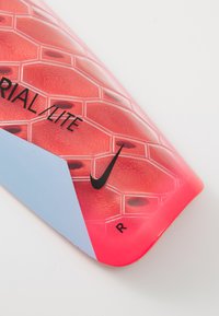 Nike jalgpalli sääreülesanne, millel on kuhexagonaalne muster, peamiselt punane siniste aktsentidega, tekstureeritud pinnaga ja julge musta brändimisega.