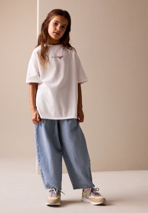 Junges Mädchen mit langen braunen Haaren trägt ein weißes, übergroßes Cars-T-Shirt, blaue und weiße gestreifte weite Hosen und bunte Turnschuhe, steht indoor.