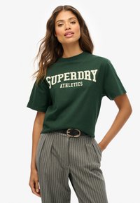 Camiseta de algodón verde con la inscripción "SUPERDRY ATHLETICS" en letras color crema, cuello redondo, mangas cortas, combinada con pantalones grises a rayas y un cinturón negro.