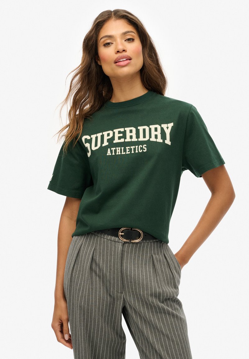 Camiseta de algodón verde con la inscripción "SUPERDRY ATHLETICS" en letras color crema, cuello redondo, mangas cortas, combinada con pantalones grises a rayas y un cinturón negro.