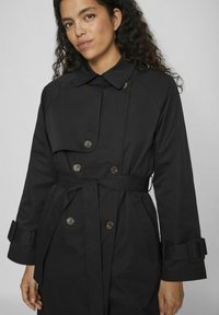 Palton negru cu nasturi dubli, guler lat, talie cu curea și manșete ajustabile. Material neted cu un design elegant.