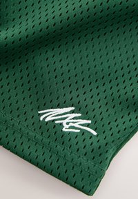 Grüner Maschenstoff mit diamantförmigen Perforationen, mit weißem, in kursiver Schrift gesticktem Logo in der unteren rechten Ecke.