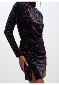 Femme portant une robe en velours noir ajustée avec des motifs floraux colorés et cachemire, manches longues, taille froncée et une fente à l'avant.