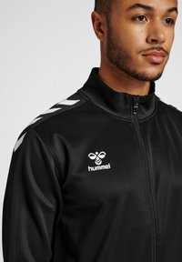 Veste à fermeture éclair noire en tissu lisse ; dotée de motifs en chevrons blancs sur les épaules et d'un logo sur la poitrine. Design près du corps.
