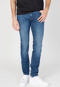 Jeans in denim blu chiaro con una vestibilità slim, dotati di cinque tasche e dettagli di cucitura discreti. Abbinati a sneaker bianche, su uno sfondo neutro.