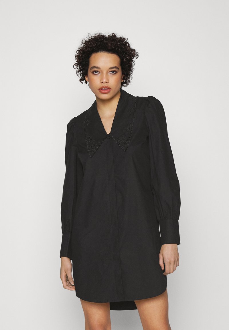Vestido preto com um colarinho grande e ornamentado. Apresenta mangas bufantes e uma frente com botões. O tecido é liso com um ligeiro brilho.