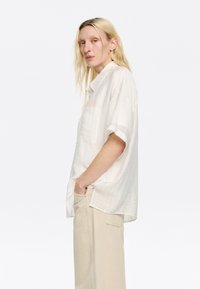 Camicia chiara a maniche corte realizzata in tessuto testurizzato con taschino, abbinata a pantaloni beige. Ha una vestibilità ampia e rilassata.