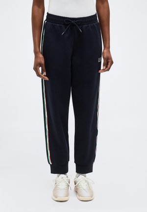 Personne portant un jogging noir avec des bandes latérales rouge, blanc et vert et des baskets blanches, debout devant un fond uni.