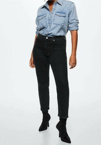 Jean noir taille haute, coupe slim et longueur cheville, associé à une chemise en denim clair à boutons et des bottines à talons noires.