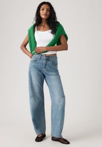 Γυναίκα με μακριά σκουρόχρωμα μαλλιά που φορεί λευκή φανέλα, πράσινο πουλόβερ περασμένο στους ώμους, ανοιχτό μπλε τζιν με φαρδιά πόδια και μαύρα παπούτσια με πλέγμα.