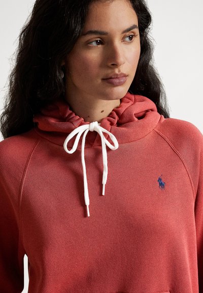 Koralfarget hettegenser med snor i halsen, ribbet tekstur og en liten blå logo på brystet. Har en avslappet passform og mykt materiale.