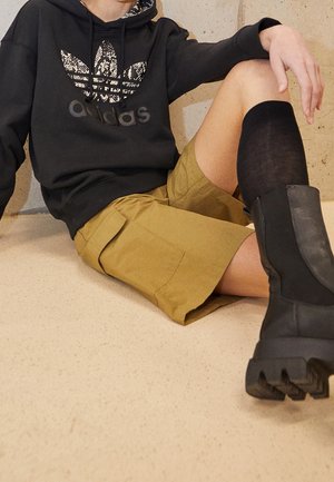 Osoba ubrana w czarną bluzę z kapturem Adidas, khaki krótkie spodnie typu cargo, czarne skarpety do kolan i czarne masywne buty, siedzi na podłodze.