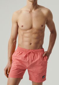 Koralowe szorty pływackie wykonane z lekkiego materiału, z elastycznym paskiem w talii i sznurkiem do regulacji, bocznymi kieszeniami oraz małą czarną naszywką logo.