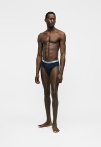 Ateetiline mees paljajalu, kandes musta aluspükse helesinise vöökohaga, millel on bränd "Emporio Armani", valgel taustal.