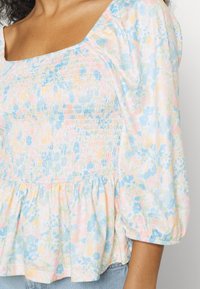 Blusa floral leve com decote quadrado, cores pastel, corpo franzido e mangas bufantes. O tecido apresenta um padrão texturado e delicado.