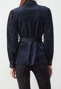 Navy blue corduroy blouse with a belted waist, puffed long sleeves, and buttoned cuffs, featuring a back pleat detail for shape.