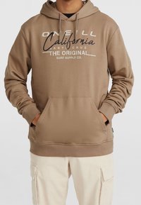 Personne portant un sweat à capuche beige avec poche avant et texte "O'Neill California Santa Cruz The Original Surf Supply Co." ainsi qu'un pantalon cargo beige clair.