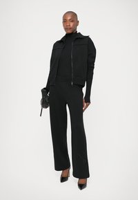 Veste noire à zip avec des manches texturées, associée à un col roulé noir ajusté et un pantalon large avec des accents de clous. Des talons noirs complètent le look.