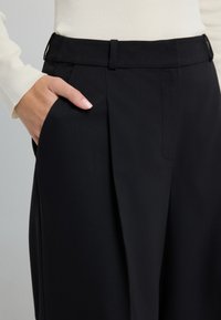 Zwarte hooggetailleerde broek van een gladde stof met zijzakken, geplooid ontwerp en ceintuurlussen. De tailleband is plat en op maat gemaakt.