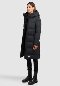 Čierny prešívaný kabát s kapucňou, so zipsovým a gombíkovým zapínaním, dvoma bočnými vreckami a logovou značkou na leme. Gumené detaily na čižmách.