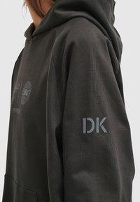 Czarna bluza z kapturem z przednią kieszenią i teksturowanym materiałem. Na lewym rękawie widnieją litery "DK" w jasnoszarym kolorze. Minimalistyczny design.