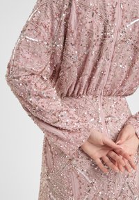 Roze paillettenjurk met ingewikkelde kraalversieringen en een gestructureerd bloemenpatroon; heeft lange mouwen en een aangesloten taille-detail.