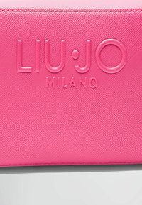 Růžový texturovaný syntetický materiál s embosovaným nápisem "LIU·JO MILANO", s hladkými akcenty a precizním prošíváním kolem okrajů.