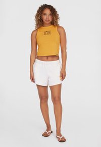 Yellow cropped tank top with "WOMEN OF THE WAVE" text, paired with white shorts featuring a small graphic. Model wears white flip-flops.