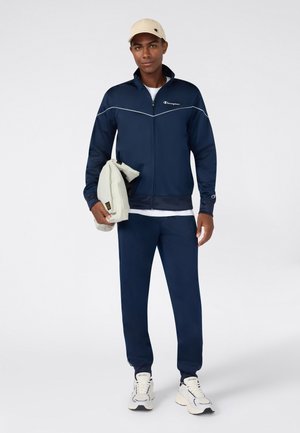 Hombre vestido con chándal azul marino y gorra beige, sosteniendo una chaqueta de color claro, de pie sobre un fondo blanco liso.