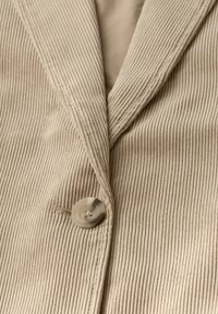 Beżowa kurtka z sztruksu z szerokimi klapami i dużym, okrągłym guzikiem z przodu.