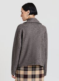 Cardigan in morbido tessuto marrone con collo pieghevole, vestibilità ampia e trama strutturata. Presenta un orlo posteriore leggermente corto ed è indossato sopra a una gonna a quadri.