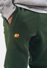 Pantaloni da jogging verdi in morbido tessuto con una tasca laterale, caratterizzati da un piccolo elefante giallo ricamato sul lato sinistro in basso.