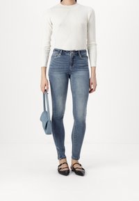 Jeans de mezclilla azul ajustados con una cintura alta, combinados con una blusa blanca de mangas largas con canalé. Un pequeño bolso azul y unos zapatos planos negros de punta completan el atuendo.