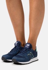 Legs wearing navy blue New Balance 500 sneakers with white soles, black outsoles, and light green accents on a white background.