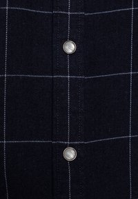 Mörkblå rutigt skjorta gjord av tyg med en mjuk textur; har silverfärgade knappar och vit sömnad i ett rutmönster.