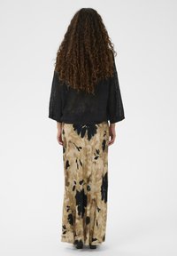 Sieviete ar gariem, lokainiem matiem, tērpusies melnā teksturētā džemperī un bēšā svārkā ar lieliem melniem ziedu rakstiem, stāv ar muguru pret skatītāju.