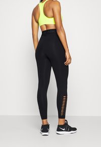 Žena v černých legínách s vysokým pasem a mřížkovými výřezy na lýtkách, s neonově žlutým sportovním podprsenkou a černými teniskami Nike, stojící na bílém pozadí.
