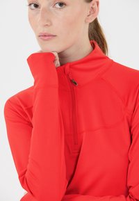 Rode atletische top met lange mouwen, voorzien van een hoge kraag en een kwartlange rits. Gladde textuur met stiksels details op de schouders.