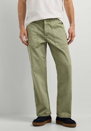 Mann trägt hellolivgrüne gerade geschnittene Hose mit großen aufgesetzten Fronttaschen und schwarzen Schuhen mit braunen Sohlen.