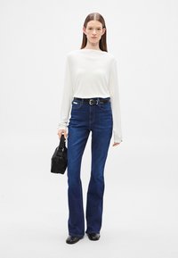 Fehér hosszú ujjú felső, sötétkék, szárán bővülő farmernadrág, fekete öv és kis fekete kézitáska. Simább anyag és klasszikus farmer textúra zsebekkel.